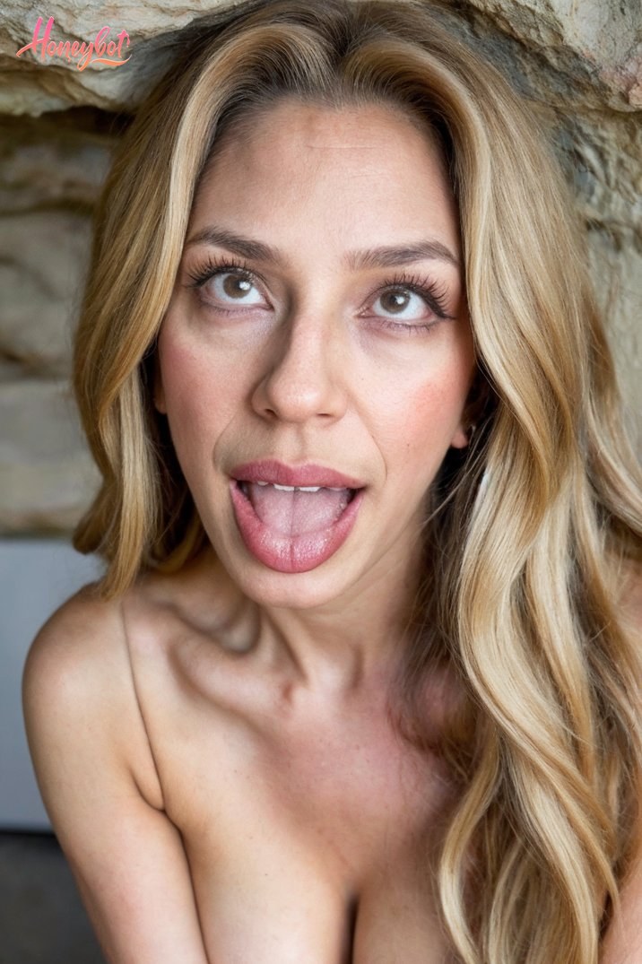 The woman in the picture has long, wavy blonde hair. She appears to be standing in front of a stone wall, which serves as a backdrop for the image. Her shoulders are bare, reveali…