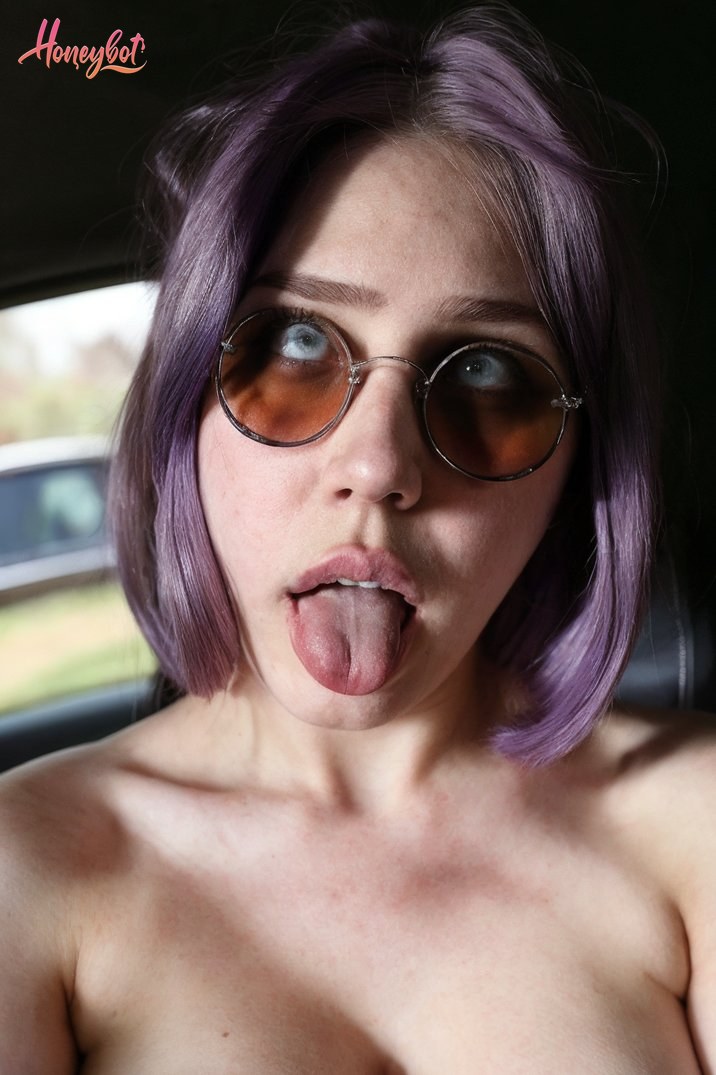 The woman in the picture has short, purple hair and is wearing round sunglasses. She appears to be inside a vehicle, as suggested by the window and interior visible in the backgro…