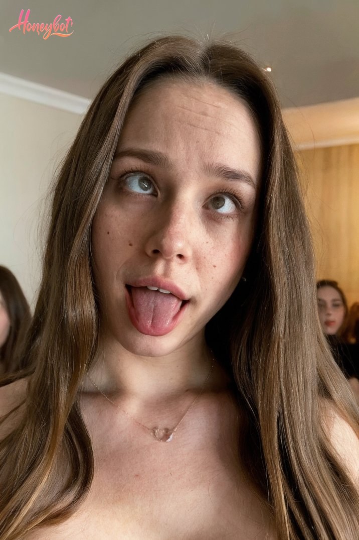 The woman in the picture has long, wavy brown hair that falls over her shoulders. She is wearing a necklace with a small pendant. Her expression appears playful, with her tongue s…