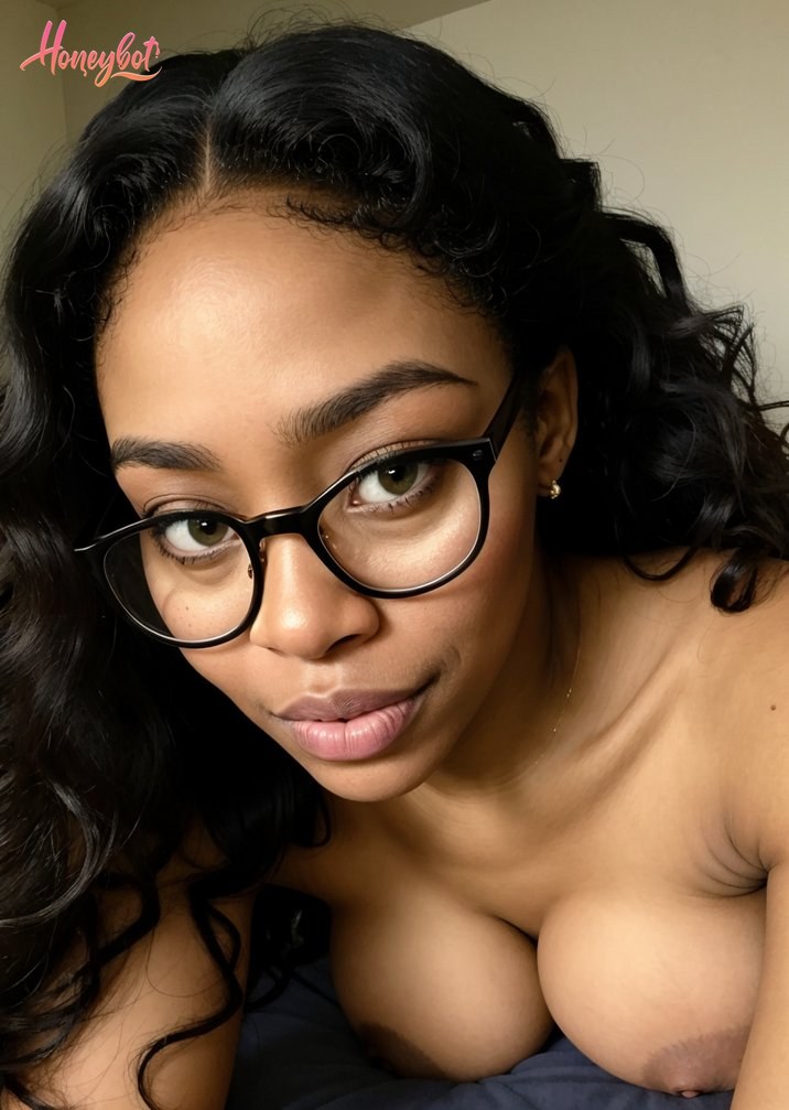 A woman with long, curly black hair and black-rimmed glasses is looking directly at the camera. She is topless, with her breasts visible. Her face is in focus, showing her eyes, n…