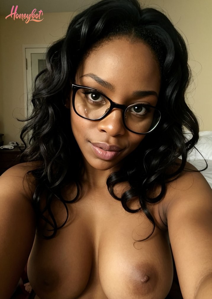 A woman with long, wavy black hair and black-rimmed glasses is topless, showing her chest and shoulders. She is looking directly at the camera with a neutral expression. Her body …